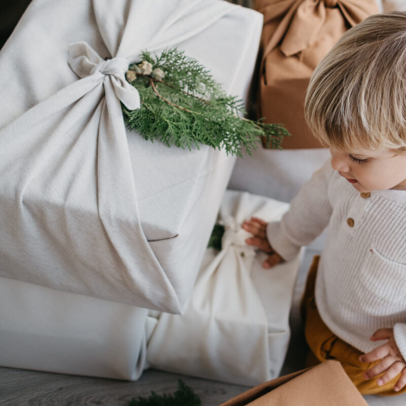 gift wrapping hacks: Cute two year old child opening presents wrapped with cloth with furoshiki technique and adorned with fir tree leaves